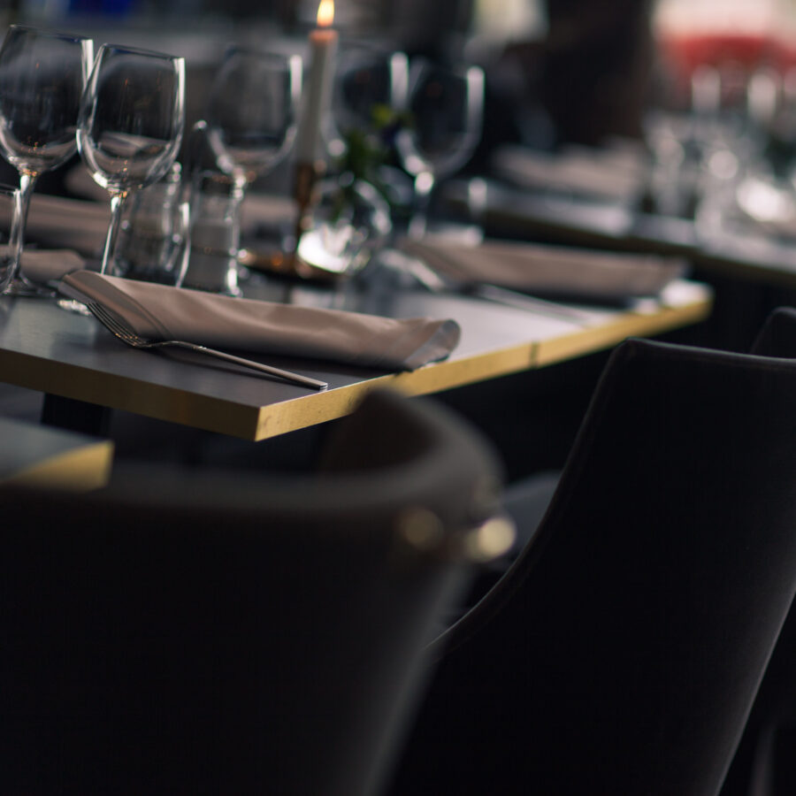 Närbild på en klädd stol och uppdukat bord i restaurangen. Bordet är vackert dukat med tallrikar, bestick, glas, servetter i linne och tända ljus i mässingsljusstakar. Stolen är klädd i ett mörkgrått tyg och har en vacker detalj av metall på ryggstödet och belysningen i restaurangen är tonad och ljusen på borden sprider ett mysigt sken. Ett exempel på hur matsalen dukas upp för kvällsgäster eller festmåltider i Bunns enda hotell och restaurang bara några mil från Jönköping och Gränna i vackra Småland.