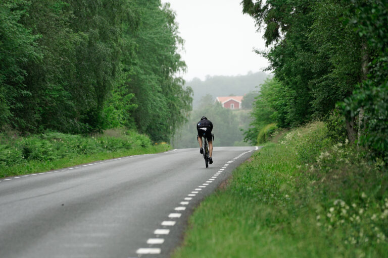 Landsvägcyklist som cyklar mot Bauergården i Småland. Han cyklar på raksträcka genom vacker lövskog och i bakgrunden skymtar ett rött hus med vita knutar.